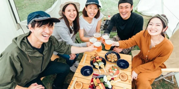 屋外で友人グループがテーブルを囲み、ビールを片手に乾杯している楽しそうな様子