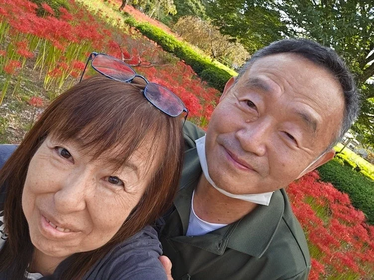 彼岸花が咲き誇る美しい背景の前で、笑顔のシニアカップルが楽しそうにセルフィーを撮っています。