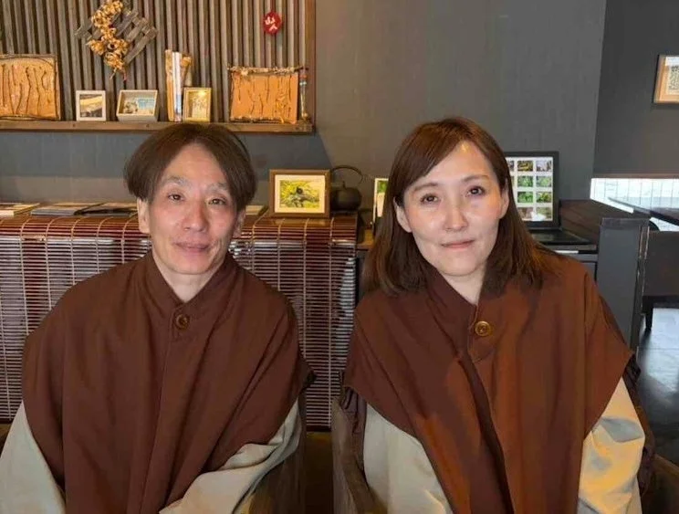 和風の室内で、茶色い上着をまとった男女が並んで座り、穏やかな笑顔を向けている様子です。