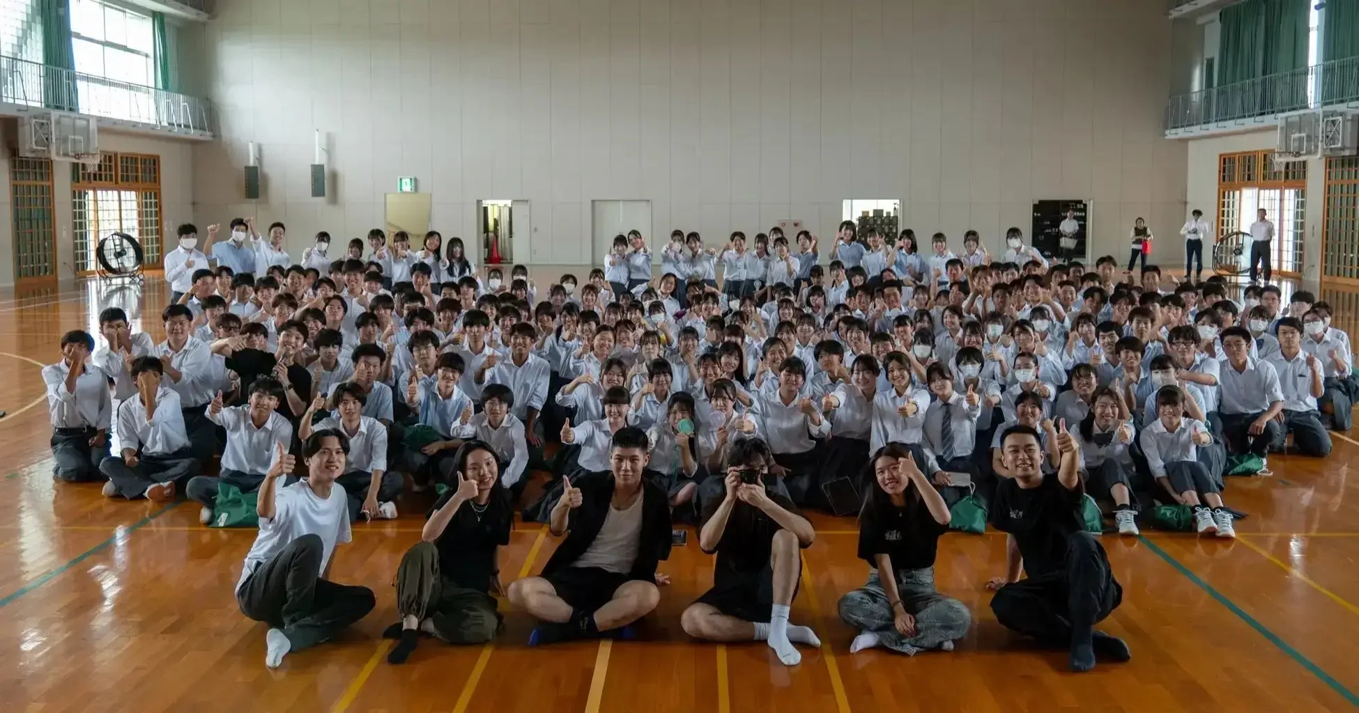 体育館で多くの学生が笑顔でサムズアップポーズをとっている集合写真