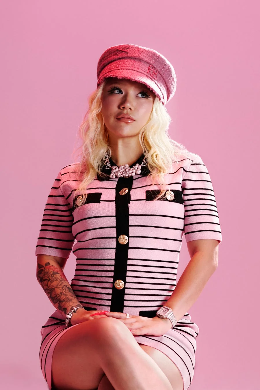 ピンクの背景で、ブロンドヘアの女性がピンクの帽子とストライプのドレスを着ています。上品なアクセサリーを身につけ、左腕にはタトゥーが見えます。スタイリッシュで魅力的なポートレートです。