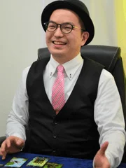 黒いハットと眼鏡をかけ、ピンクのネクタイとベストを着用した男性が笑顔を見せているポートレート